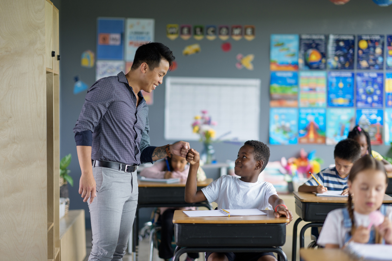A smiling teacher gives a fist bump to a smiling student sitting at a desk.