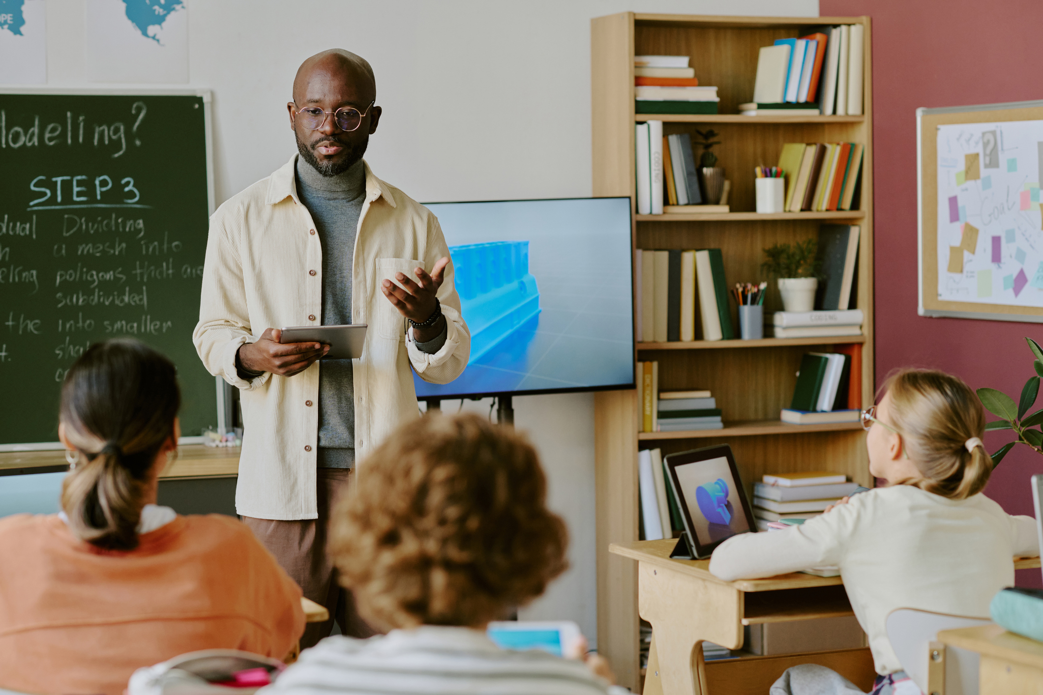 A teacher holding a tablet stands in front of students in a classroom.